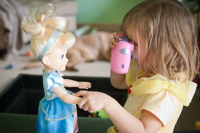 Child in yellow dress holding a pink camera. Taking a photograph of a doll in a blue dress. Photography for kids example.